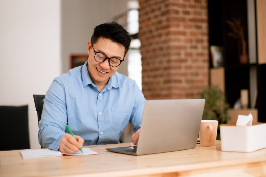 Happy asian man making notes, analyzing data for investment at workplace with laptop computer in